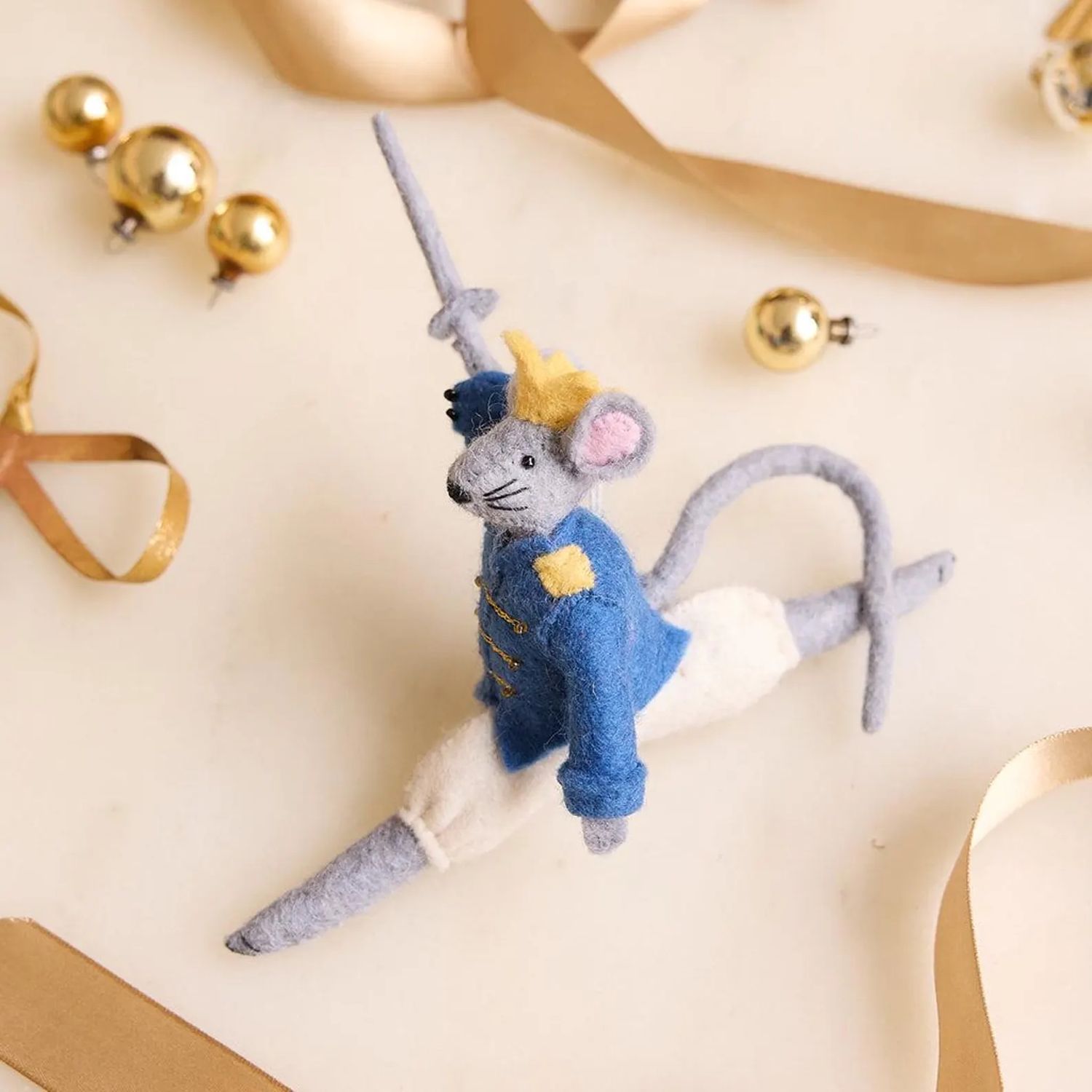 Felt mouse ornament dressed as a soldier performing a ballet pose surrounded by golden decorative objects and ribbons