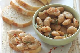 A bowl of seasoned beans garnished with herbs served alongside slices of bread and a prepared bruschetta with the beans topping