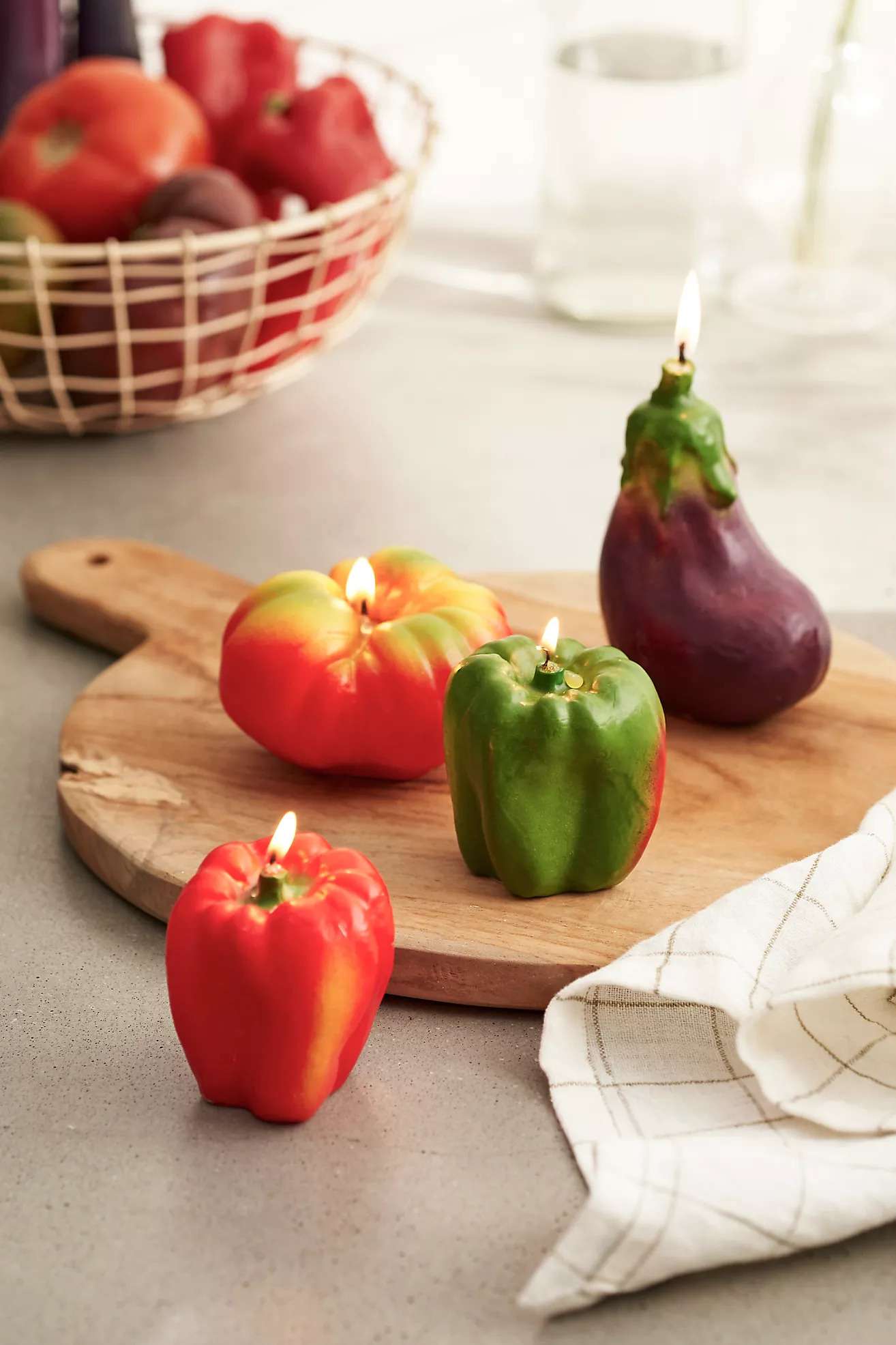 Vegetable-shaped candles on a wooden board with a towel and basket of fruits in the background