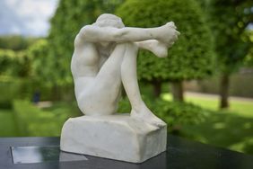 A sculpture of a seated figure hugging their knees, displayed outdoors on a pedestal