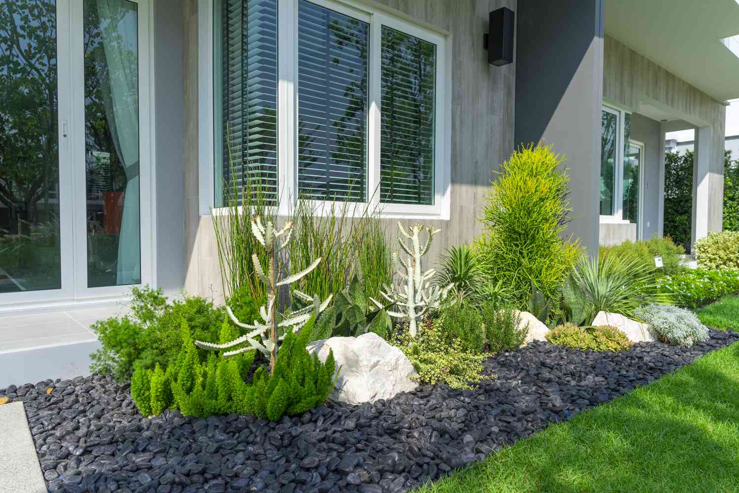 A landscaped garden area featuring a variety of plants and decorative rocks near a house with large windows