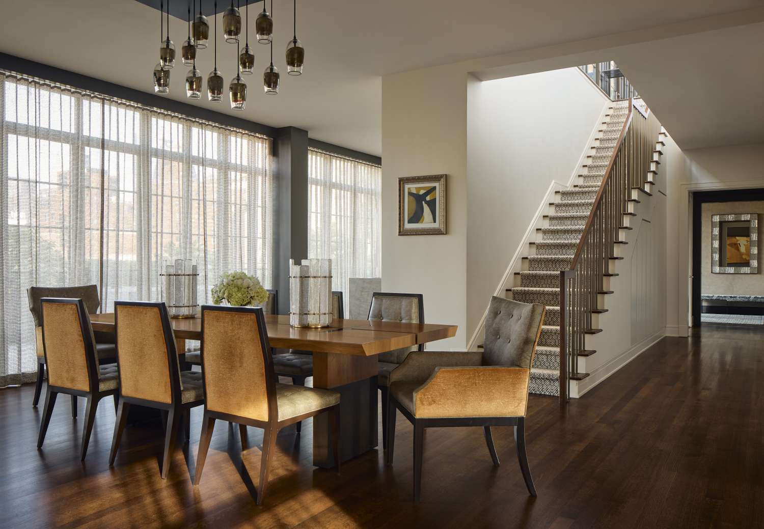 A dining room with a long wooden table chairs and sheer panel curtains adjacent to a modern staircase