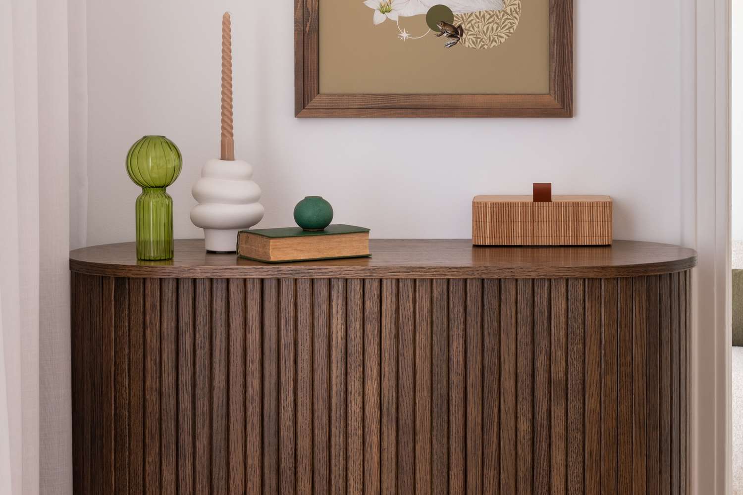 A wooden fluted sideboard topped with decorative items including a green vase a white ceramic piece with a candle and framed wall art above
