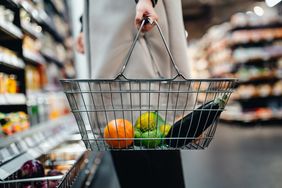 A person holding a shopping basket with fruits and a zucchini in a grocery store aisle