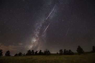 Night sky with visible Milky Way and shooting stars above a landscape with trees in the foreground