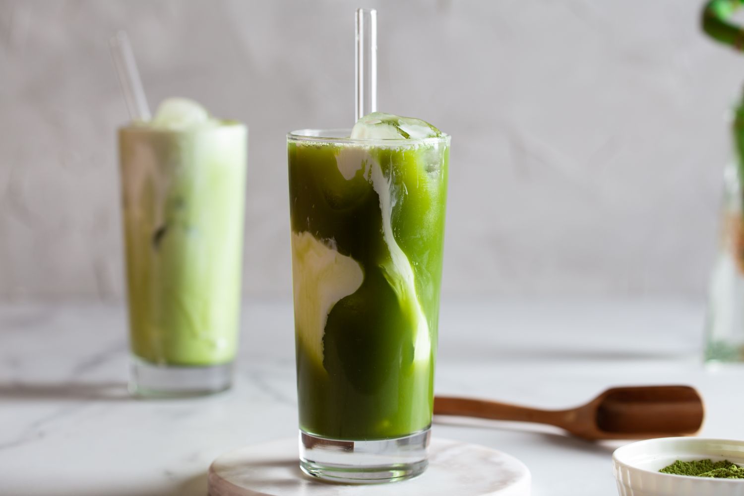 Two glasses of matcha latte with swirling milk one in the foreground and one blurred in the background on a light surface