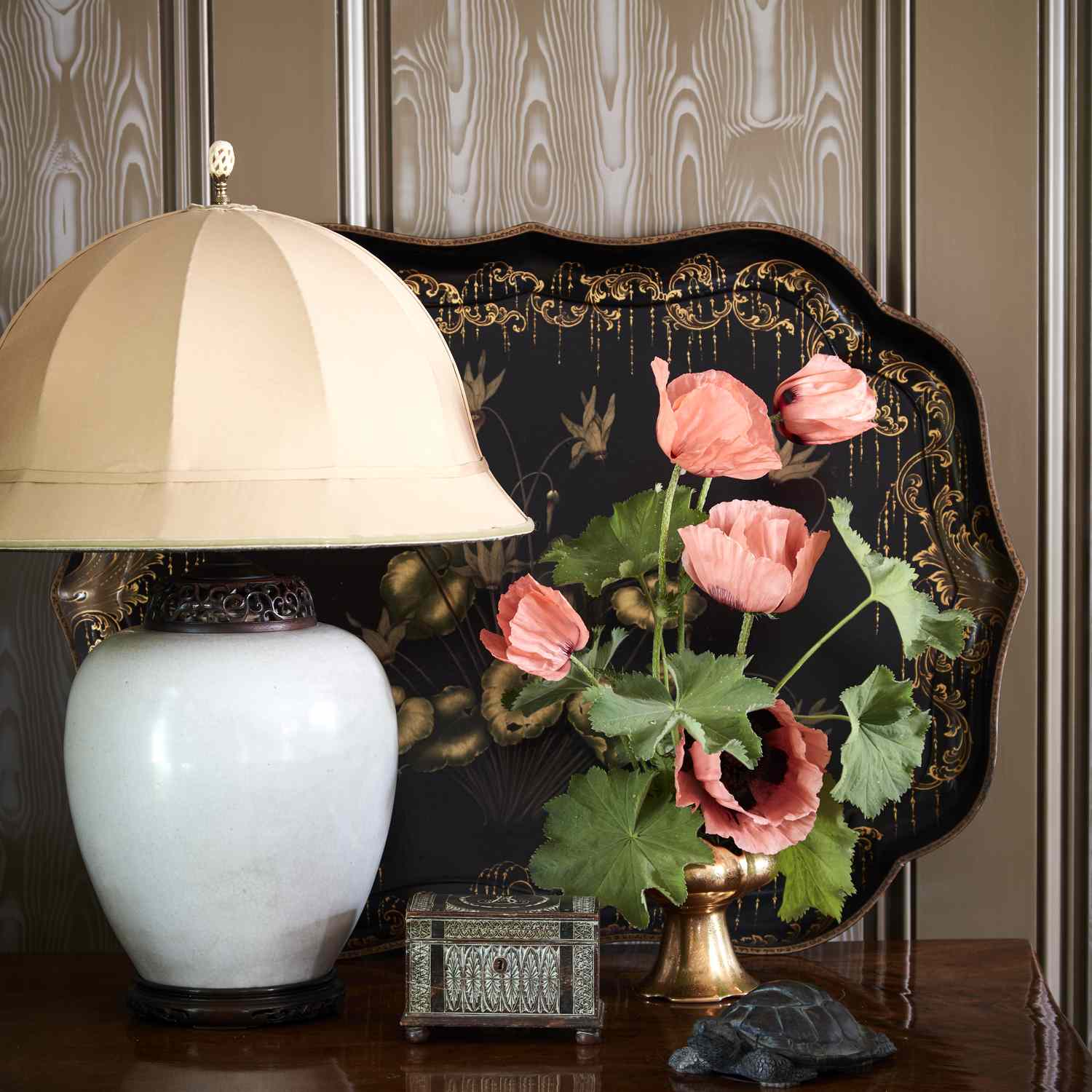 A table lamp a vase with flowers a decorative tray and small objects placed on a wooden surface