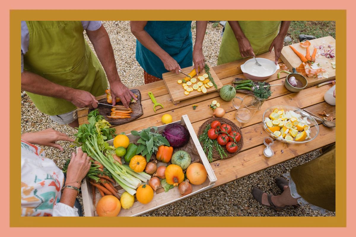 A group of people preparing vegetables on a wooden table outdoors surrounded by various fresh produce