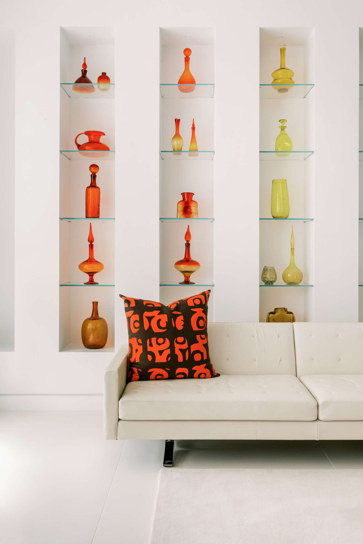 Minimalist living room by Lambrakos Studio: white leather sofa with orange-and-black patterned pillow, recessed shelves with colorful retro glassware in orange, yellow, and amber, neutral rug, white walls, and natural light create a calm, stylish space.
