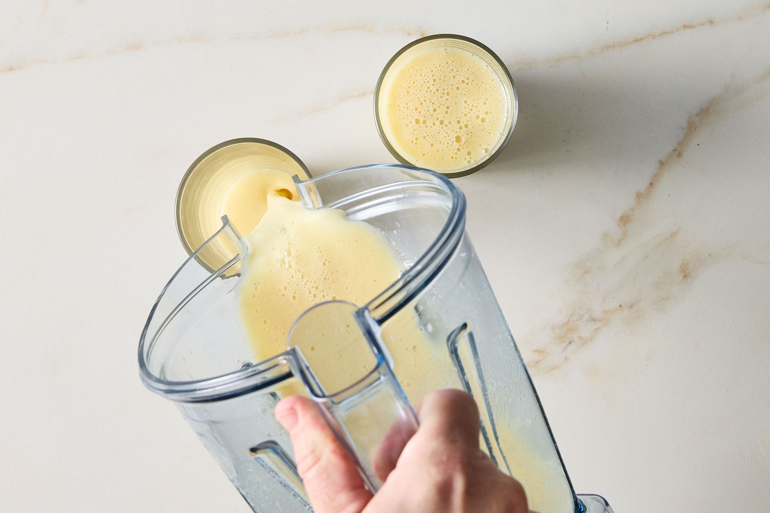 Pineapple and ginger smoothie being poured from a blender into a glass with another filled glass beside it on a countertop