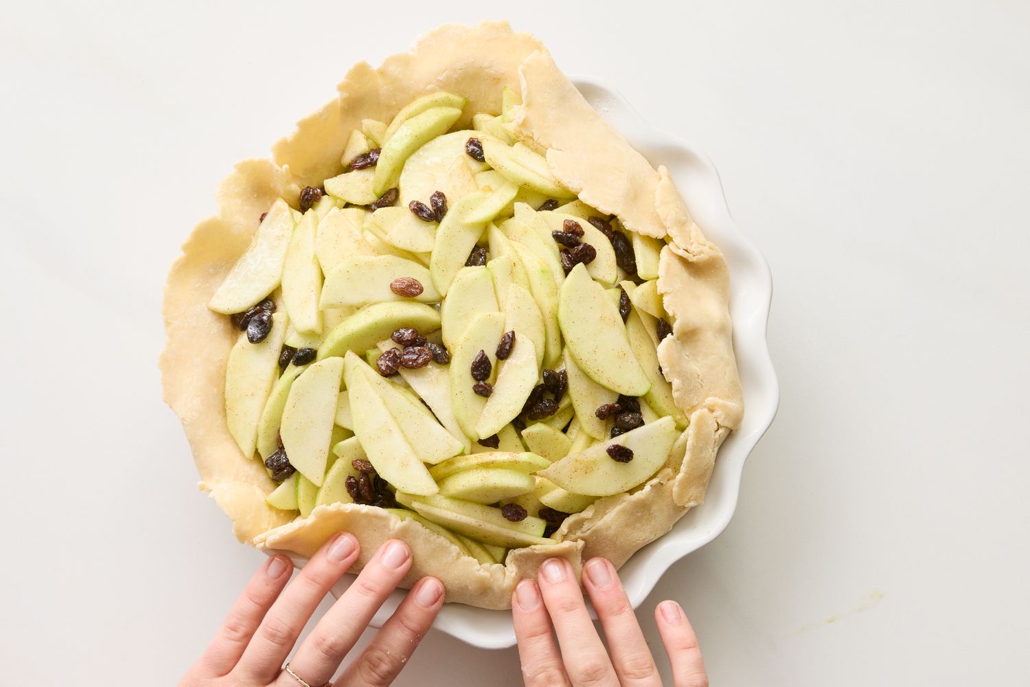 Hands arranging apple slices with raisins on a pie crust in a pie dish