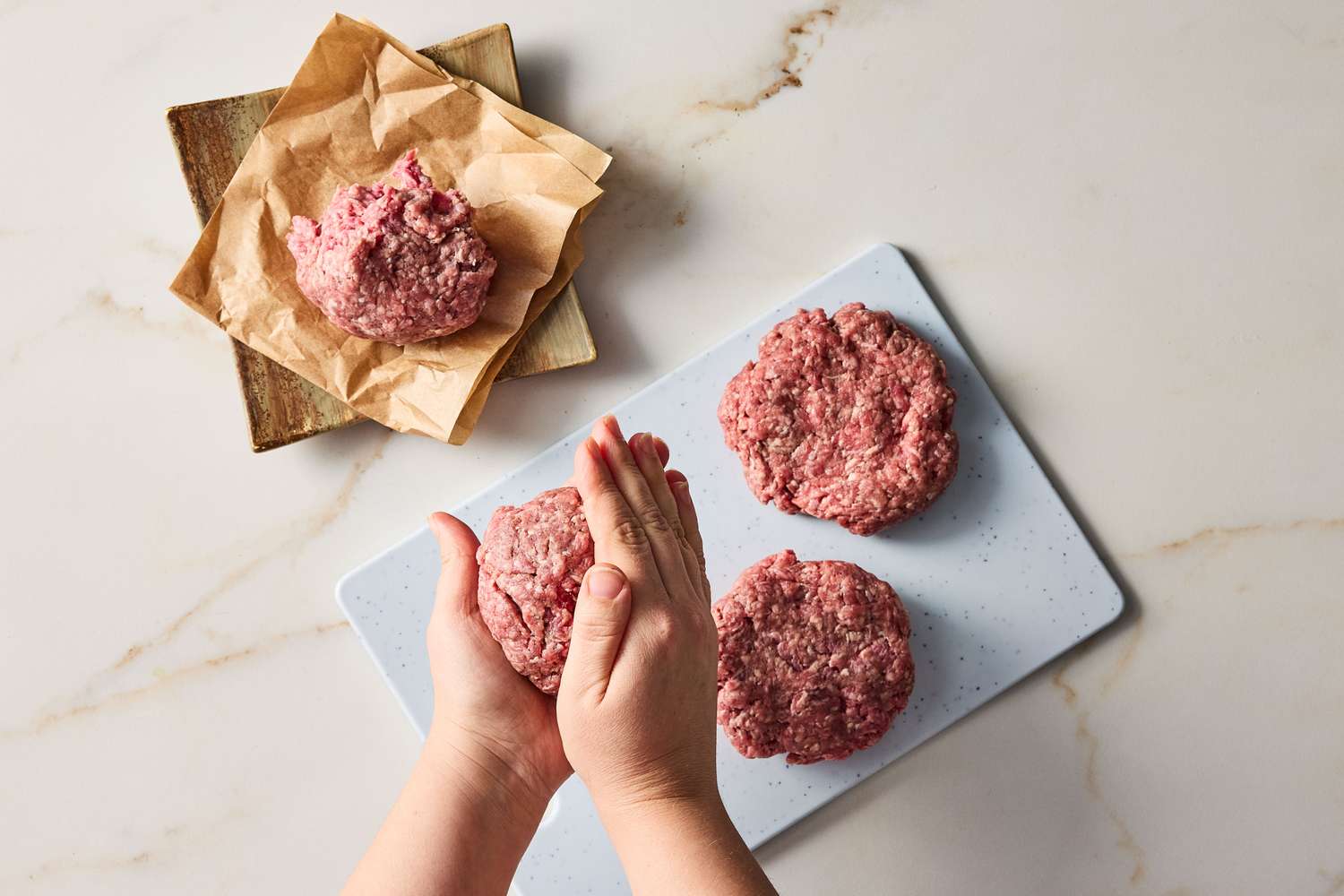 Hands shaping ground meat into patties on a cutting board with additional patties and meat portions nearby