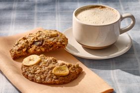 Two breakfast cookies with banana slices on a napkin beside a cup of coffee on a table