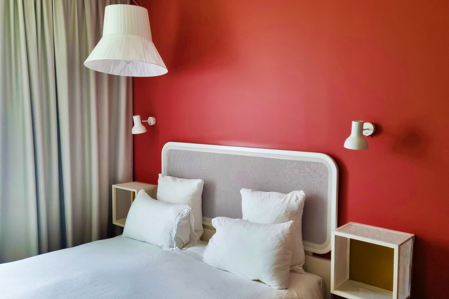 A neatly arranged bed with white linens flanked by small shelves and wallmounted lamps against a red wall background