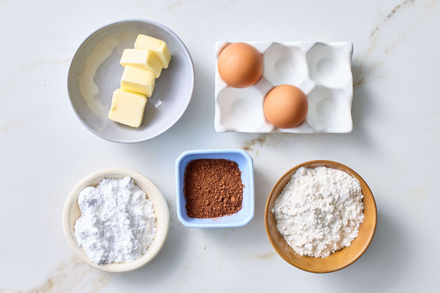 Ingredients for making a dessert, including butter, eggs, powdered sugar, cocoa powder, and flour, arranged in bowls