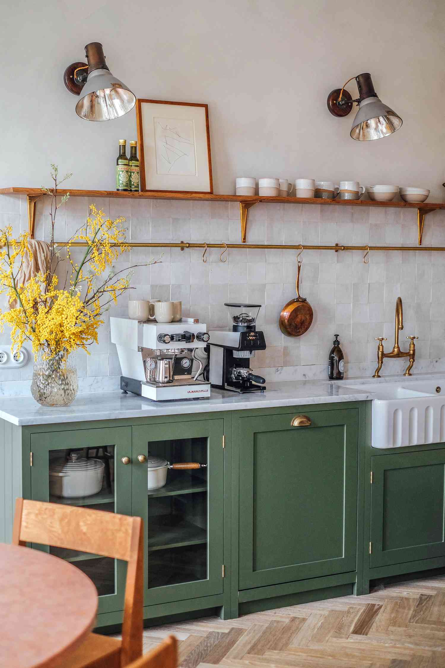 Kitchen design by Camilla Masi of Otto Tiles: deep olive cabinetry, brass hardware, farmhouse sink, and open wood shelving. A zellige tile backsplash and vintage-inspired wall sconces add warmth and texture.