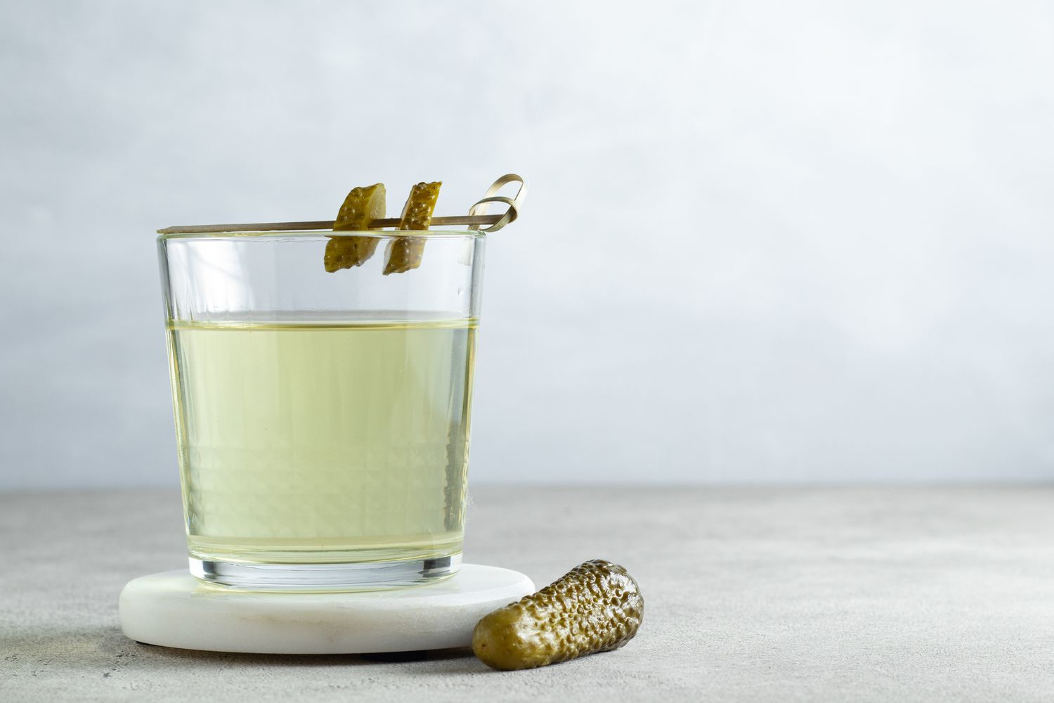 A glass filled with a lightcolored liquid garnished with pickles on a skewer and a pickle on the surface next to it