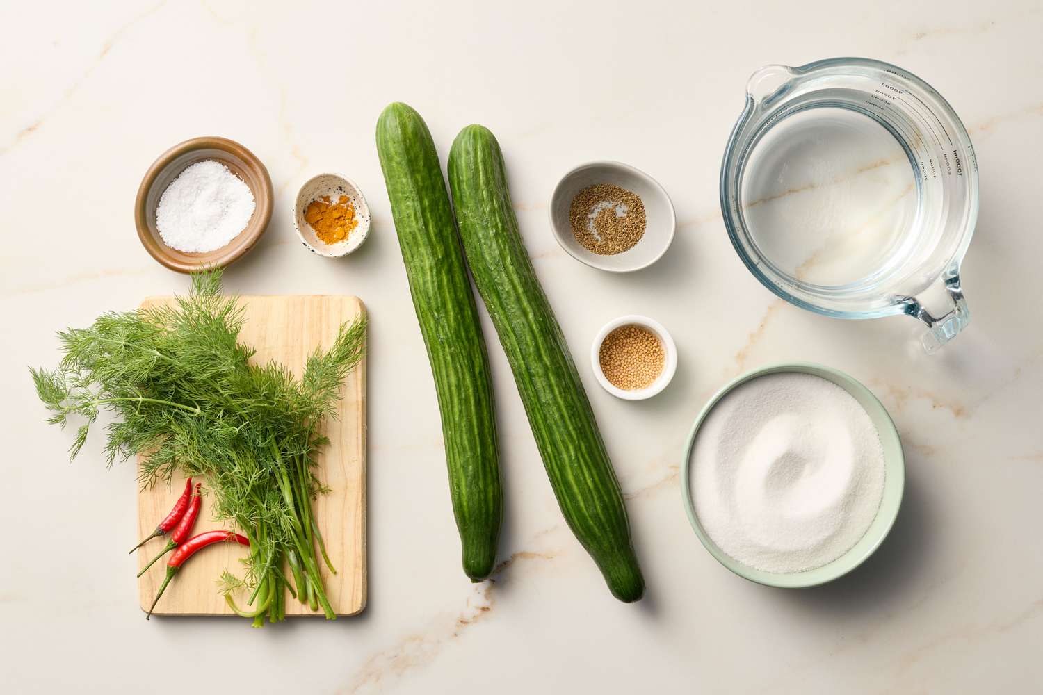 Arrangement of pickling ingredients including cucumbers, dill, chili peppers, spices, and a jar of water on a countertop