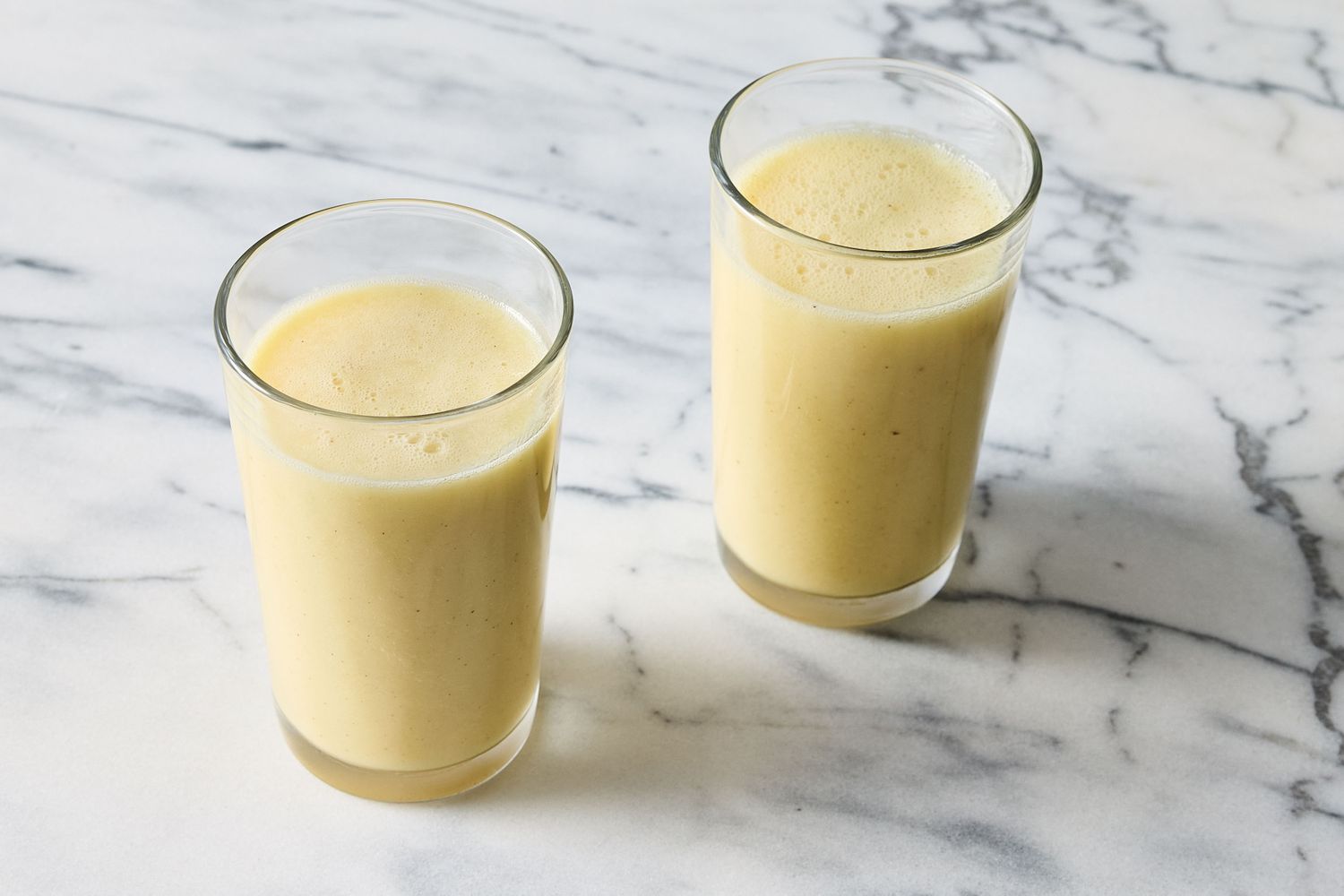 Two glasses of smoothie placed on a marble surface the drink appears creamy and is likely made with pineapple and ginger