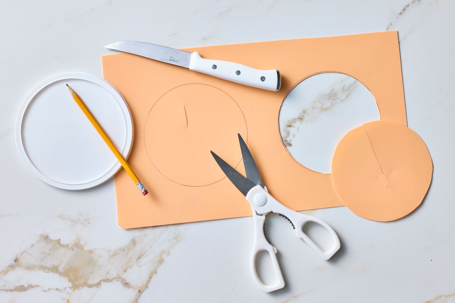 Materials for crafting including scissors, a knife, paper circles, and a pencil on a marble surface