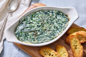 Baked spinach dip in a white casserole dish on a wooden board with slices of toasted bread
