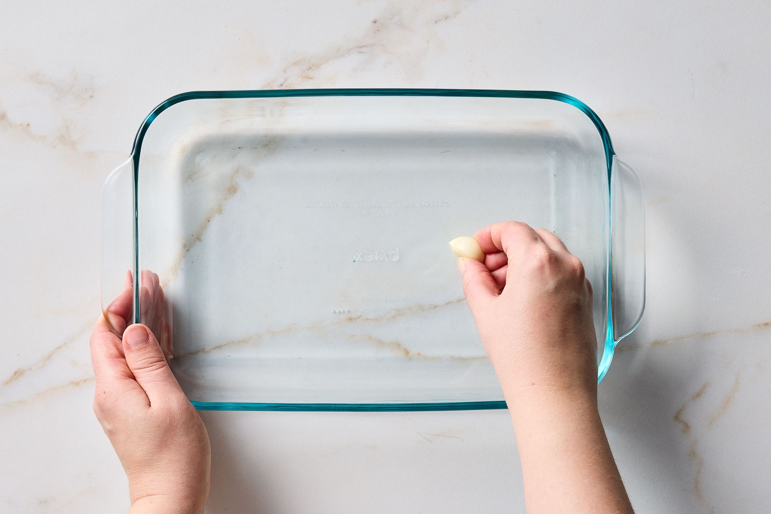 Hands rubbing a clear glass baking dish with a piece of garlic preparation for cooking
