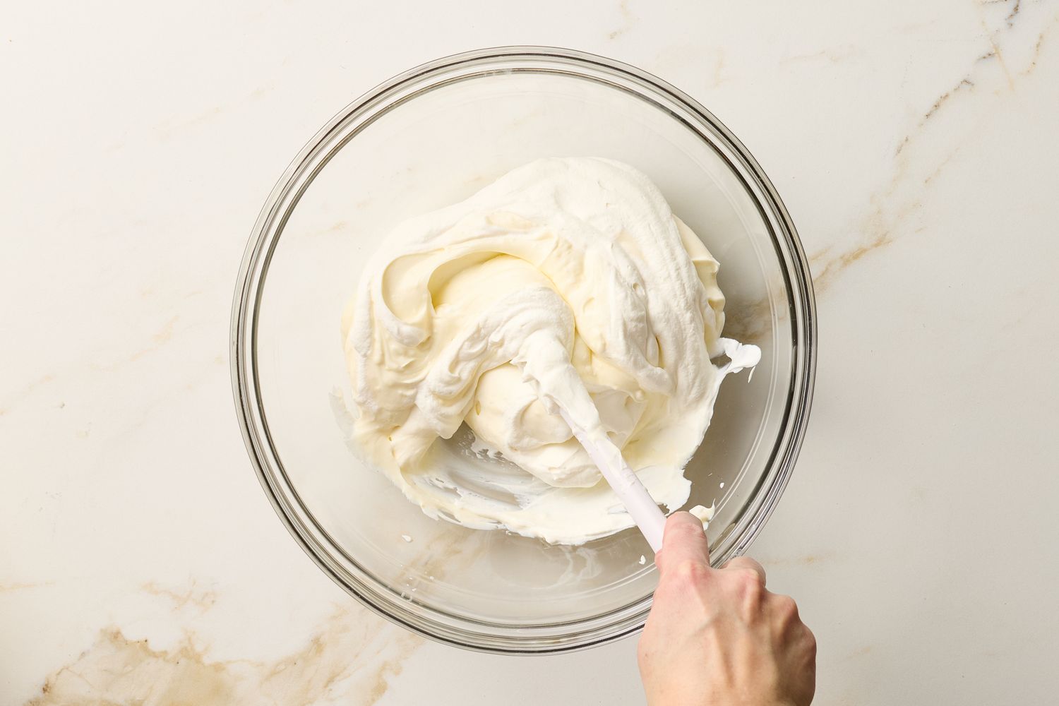 A person stirring a creamy ricotta mixture in a glass bowl using a spatula