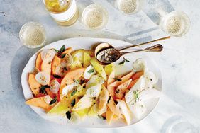 A plate of melon and cucumber salad with seasoning and a serving spoon glassware and a bottle in the background