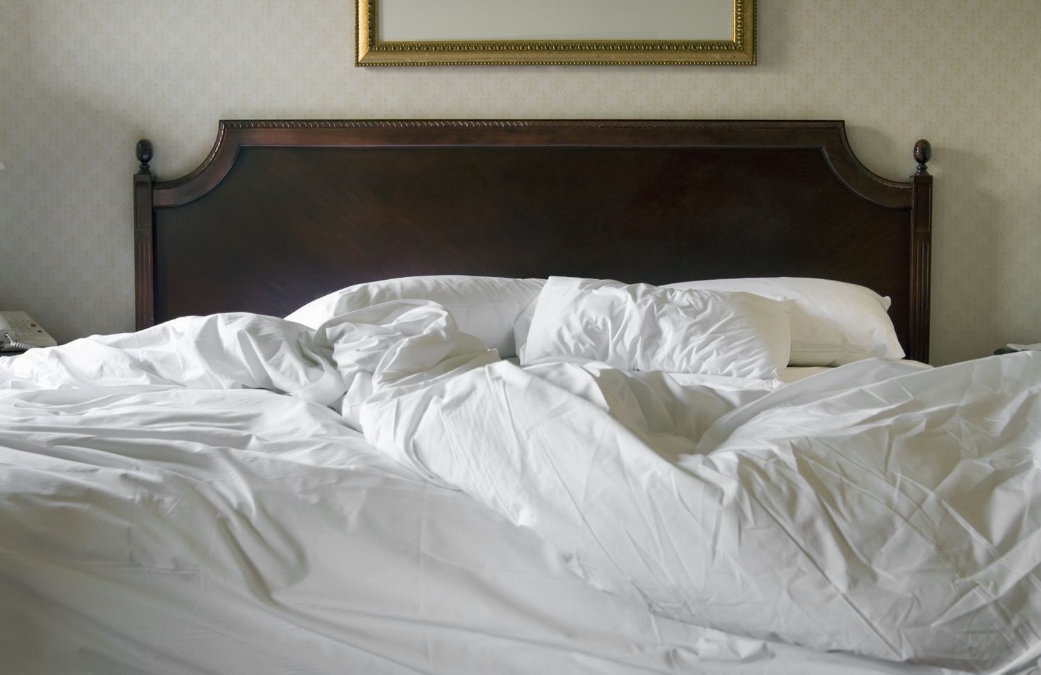 A bed with a wooden headboard and unmade white sheets and pillows in a room with partially visible decor