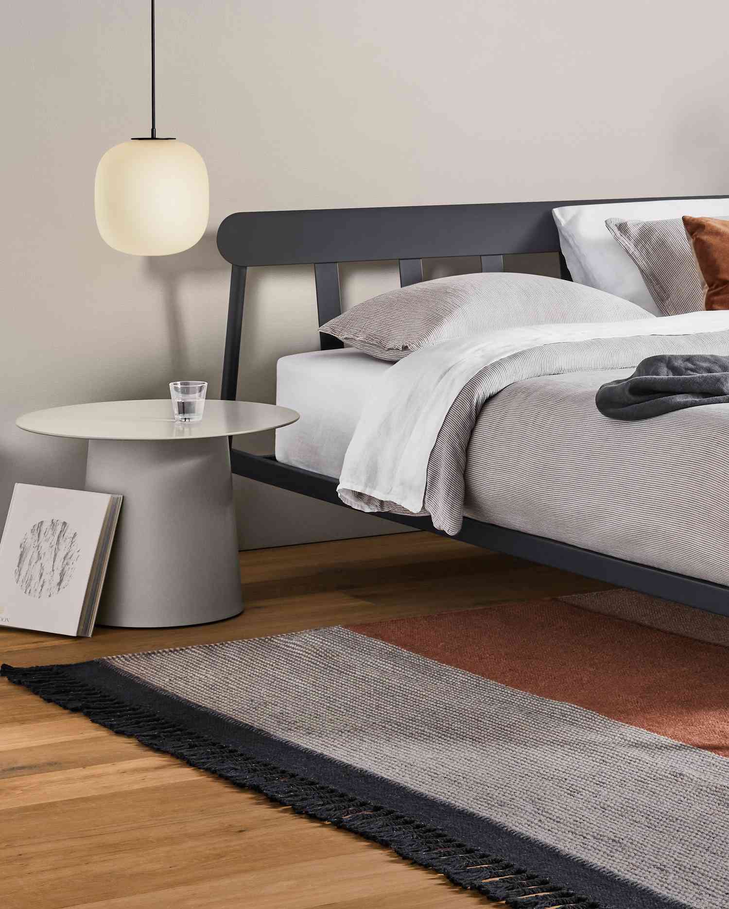 Close up shot of minimal black and grey bedroom with metal bed frame, side table, and striped hand knotted rug below. 