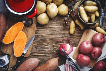 raw sweet potatoes and 3 types of white potatoes on wood surface with peeler and knives