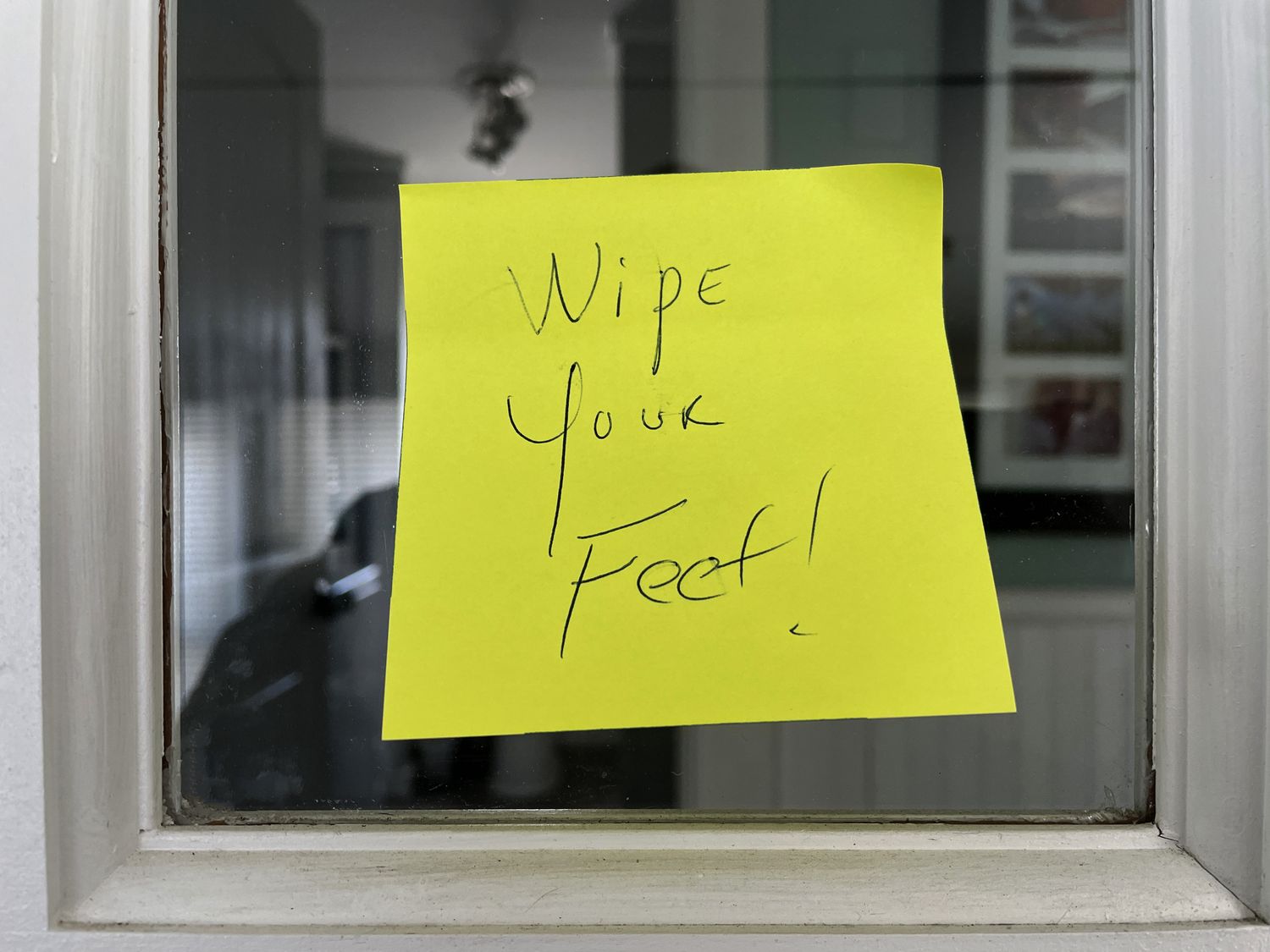 A yellow sticky note on a glass door with the message Wipe your feet written on it