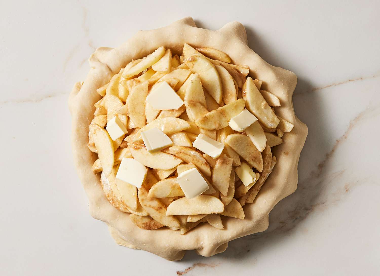 sliced apples and butter in a pie crust on a marble surface