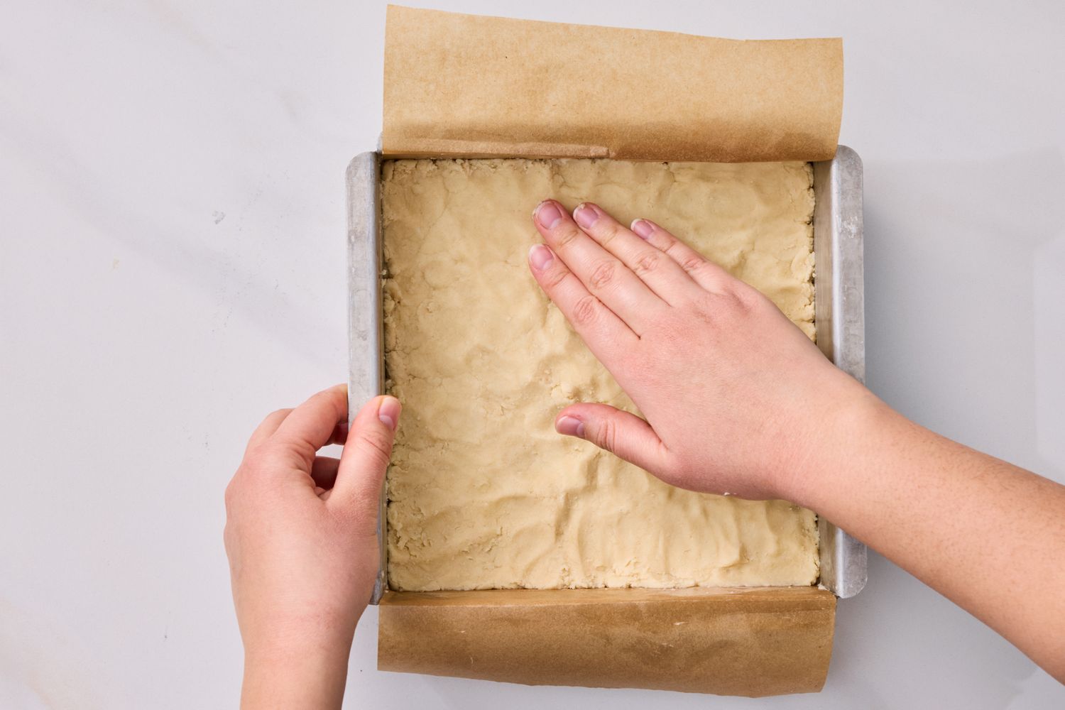 Hands pressing dough into a parchmentlined square baking pan preparing lemon squares