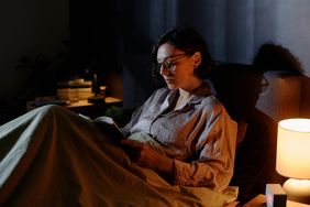 A person reading a book while sitting in a dimly lit room with a bedside lamp glowing softly