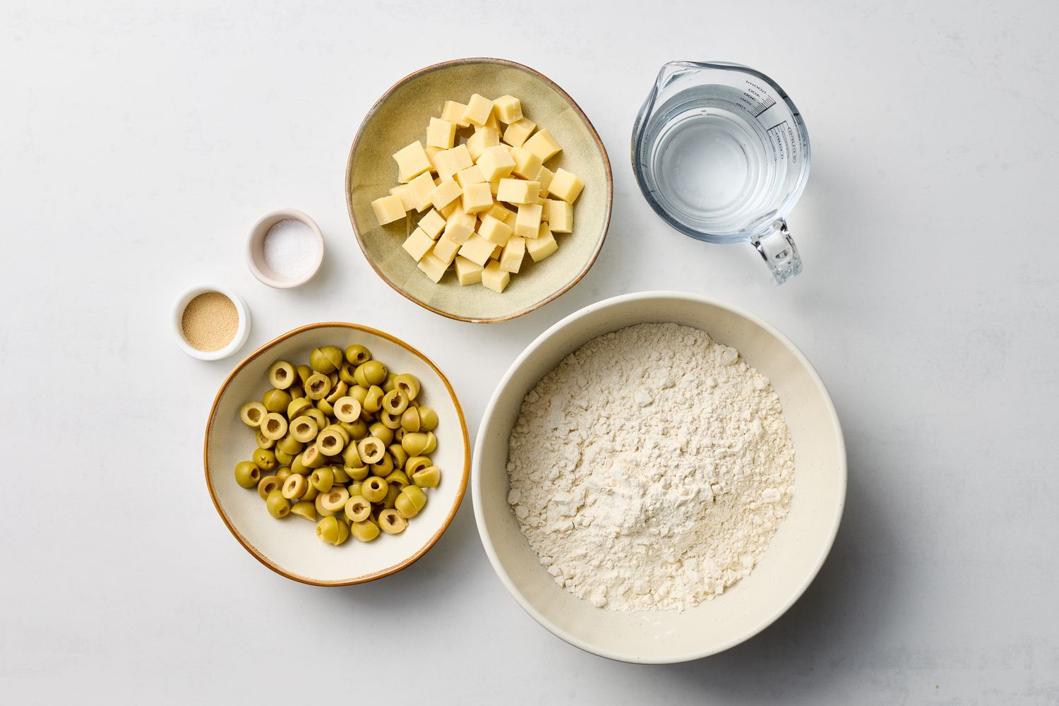 Ingredients for an olive and cheese loaf including bowls of flour chopped cheese sliced olives water and seasonings