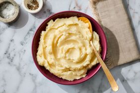 A bowl of mashed potatoes with a spoon on a marble surface next to small bowls of salt and pepper