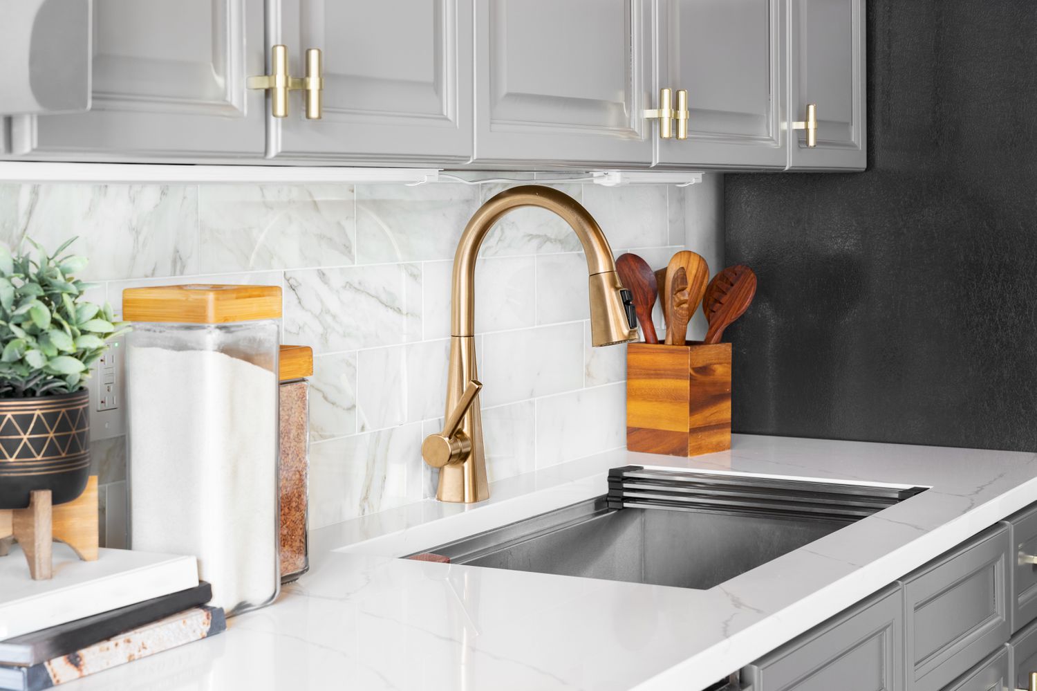 A kitchen sink detail shot with grey cabinets, a marble subway tile backsplash, and gold hardware and faucet.