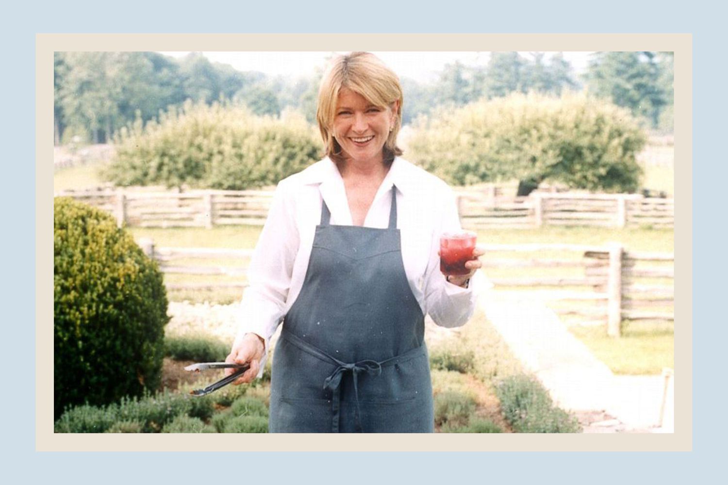 decorvow holding a drink and a pair of tongs, smiling outdoors near a garden setting