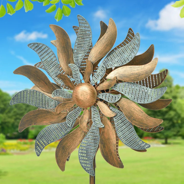 A metallic wind spinner in the shape of a flower placed in a garden setting with a grassy lawn and trees