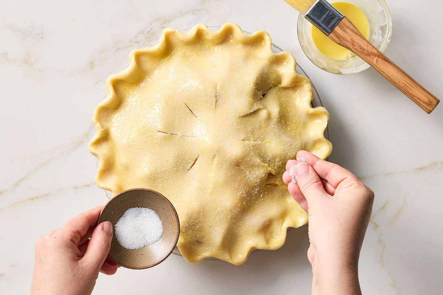 Hands sprinkling sugar on a prepared pie crust before baking with a dish of salt and a brush nearby on the counter