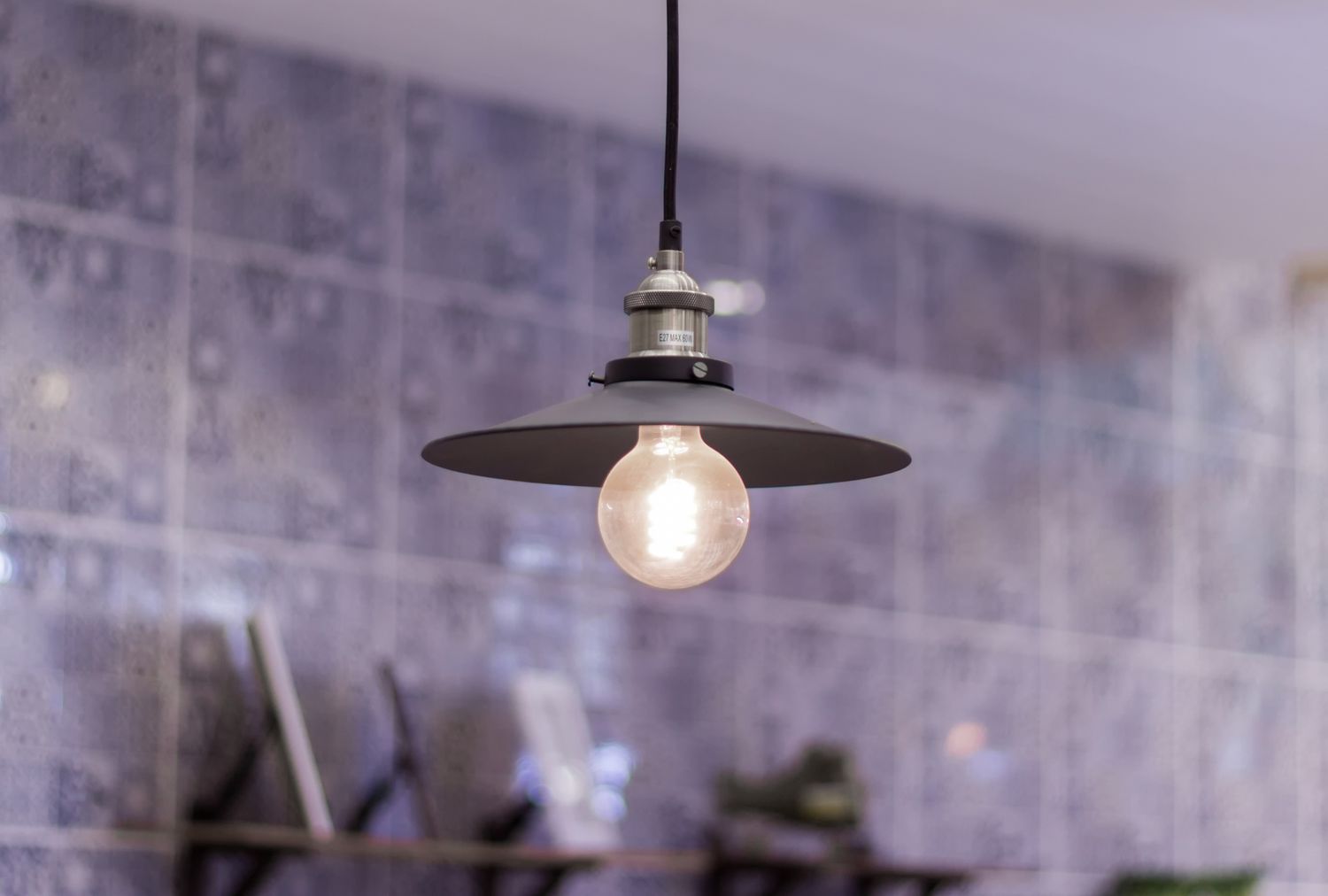 Hanging light fixture with metallic shade and exposed bulb in front of a tiled background