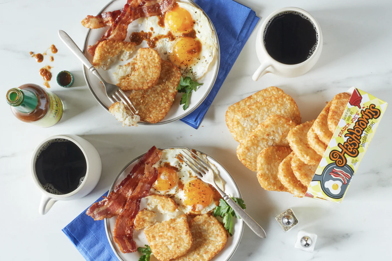 Two breakfast plates with eggs bacon hash browns coffee mugs and a box of Trader Joes hash browns shown on the side