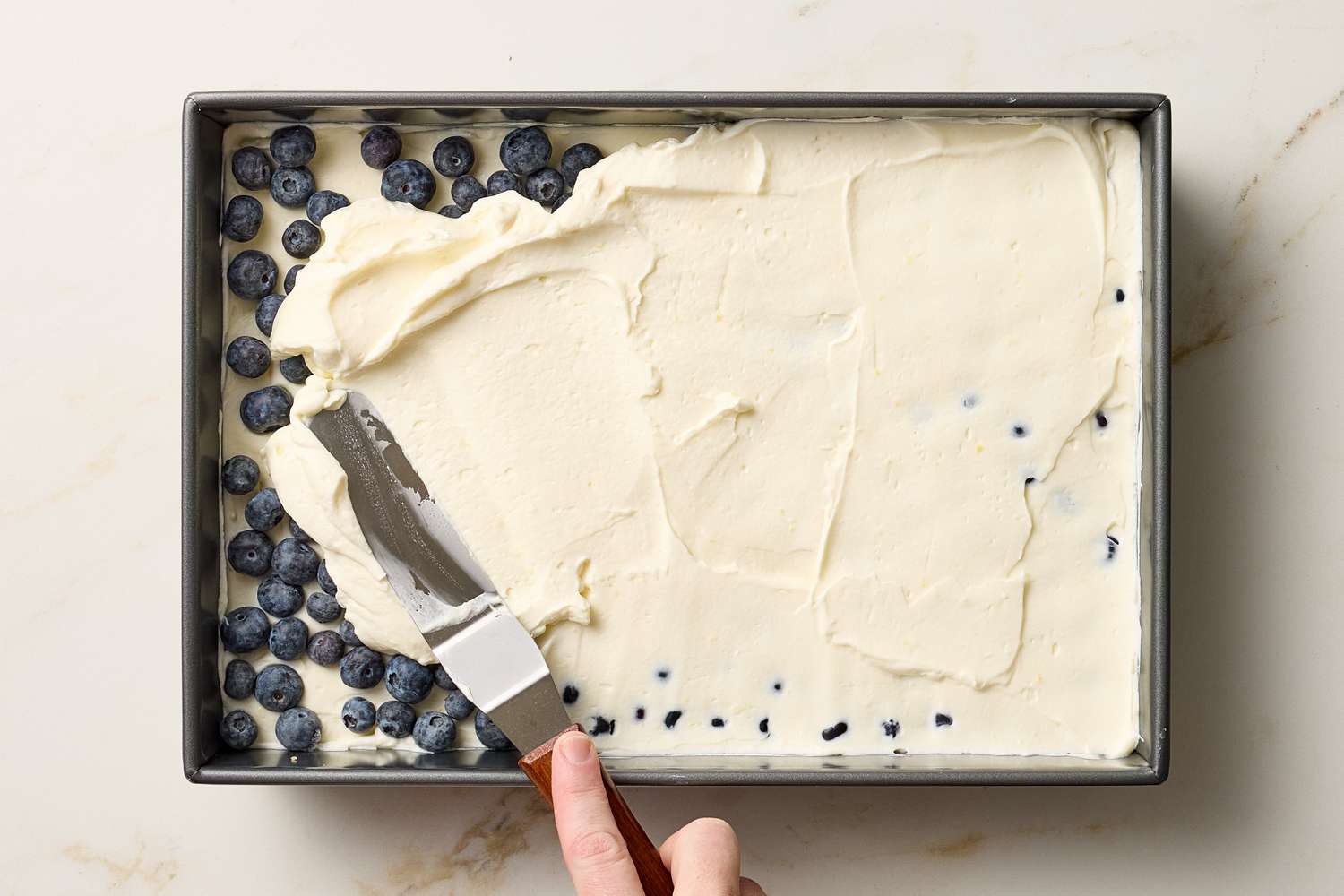 A hand spreading cream on a layered dessert with blueberries in a rectangular pan