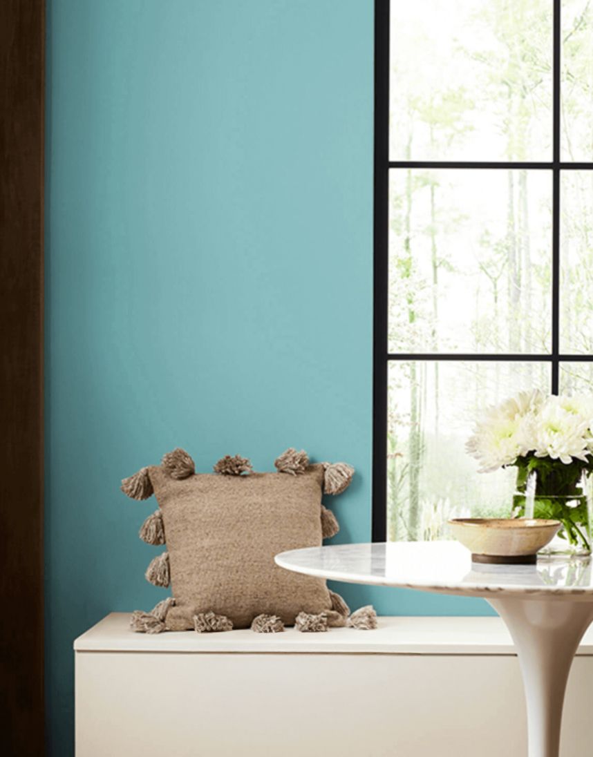A throw pillow on a bench against a teal wall beside a window and a small table with a flower arrangement