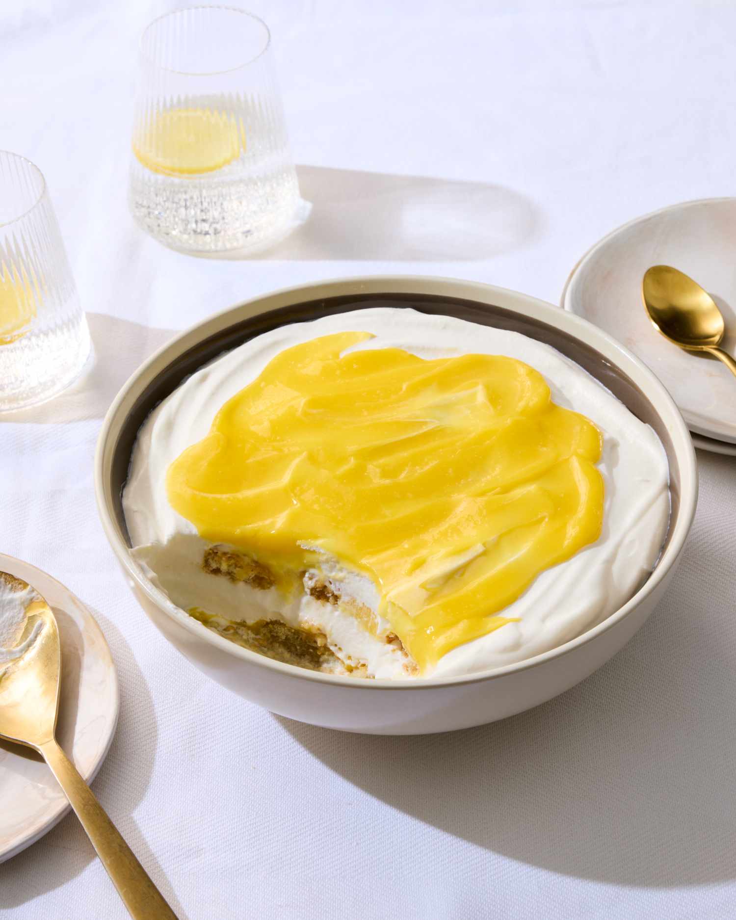 A lemon trifle dessert in a bowl with cream and lemon topping served on a table with plates spoons and glasses
