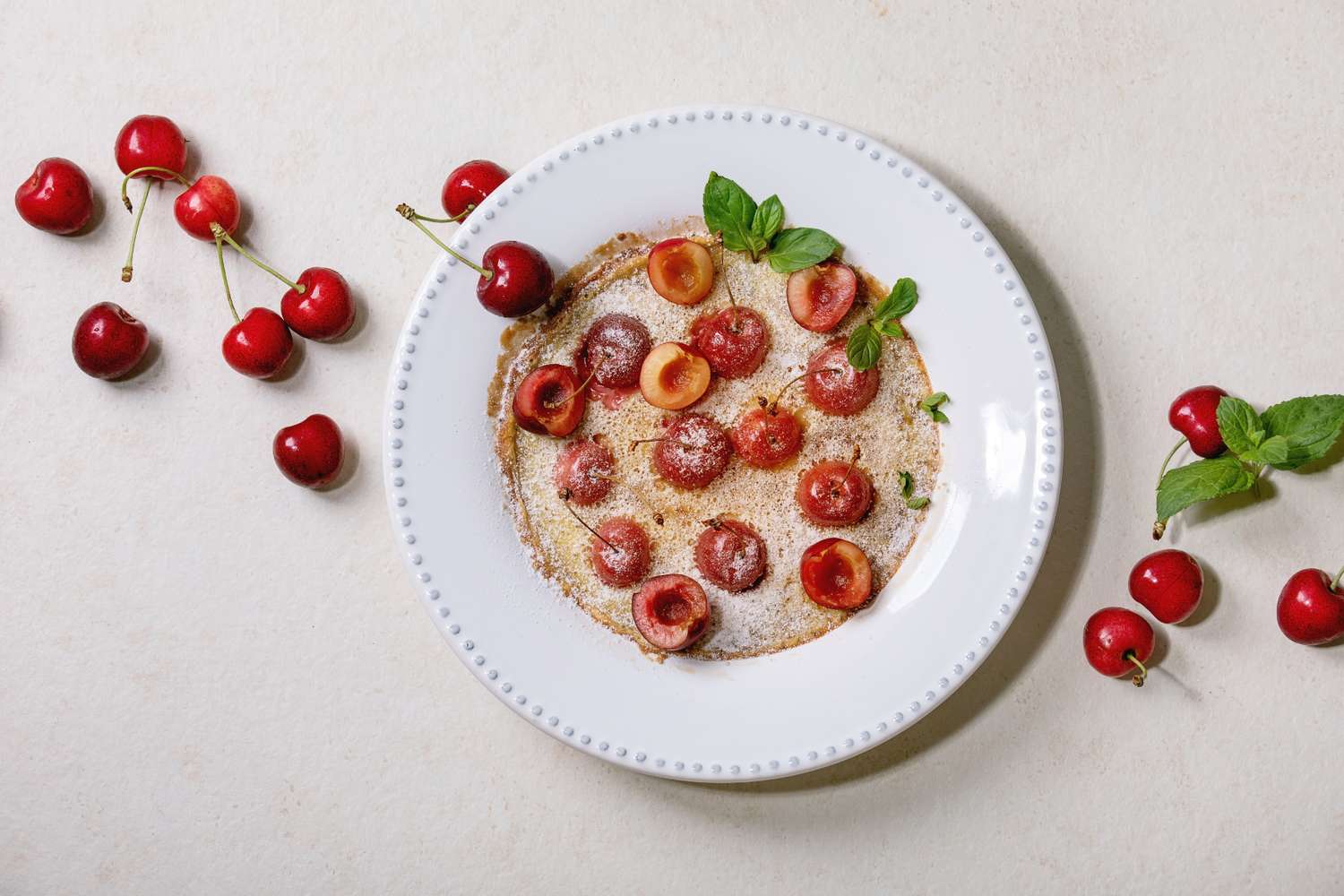 A plate with a pancake topped with cherries and garnished with mint surrounded by whole cherries and mint leaves