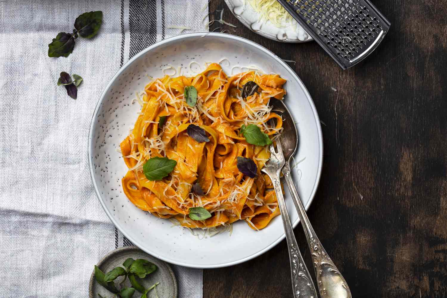 A plate of pasta garnished with herbs and accompanied by a grater and a small bowl of leaves
