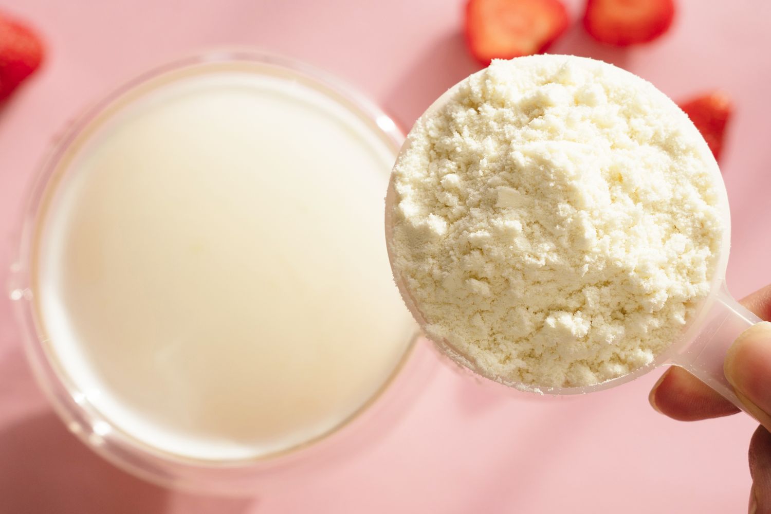 Scoop of protein powder next to a glass of liquid with strawberries on a pink background