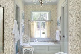 A bathroom with a patterned wallpaper a bathtub centered below a window a chair and neatly arranged towels and artwork